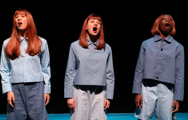 Trois femmes se tiennnent debout les bras le long du corps et chantent. Elles ont toutes la même coupe de cheveux et sont habillées en bleu.