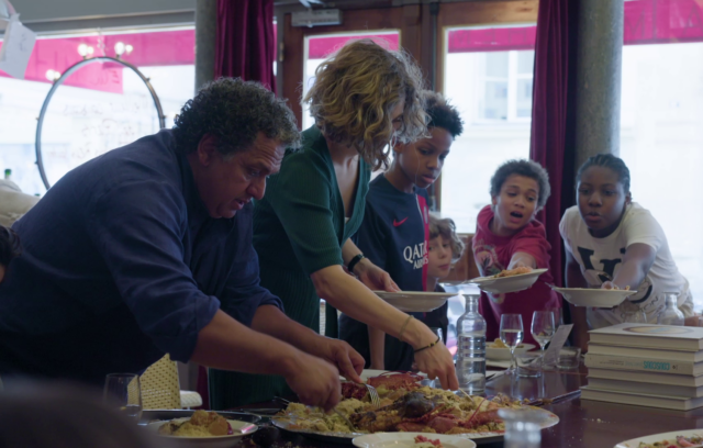 L'image provient du film Belleville Kitchen. Elle montre 2 adultes, un homme et une femme, ainsi que 3 enfants se servir d'une part de gateau sur une grande table.