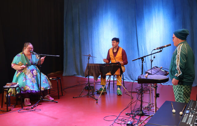 On voit 3 musiciens et musiciennes vêtus de vêtements très colorés. Iels jouent de la musique et chantent.