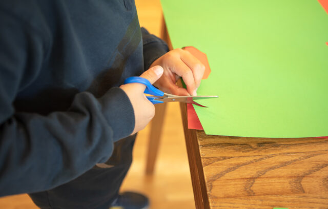 Sur l'image on voit des mains d'enfants qui découpent du papier avec une paire de ciseaux.
