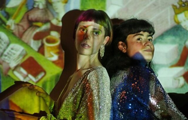 On voit les deux musiciennes du groupe BAIX. Elles sont dos à dos sur un fond coloré.