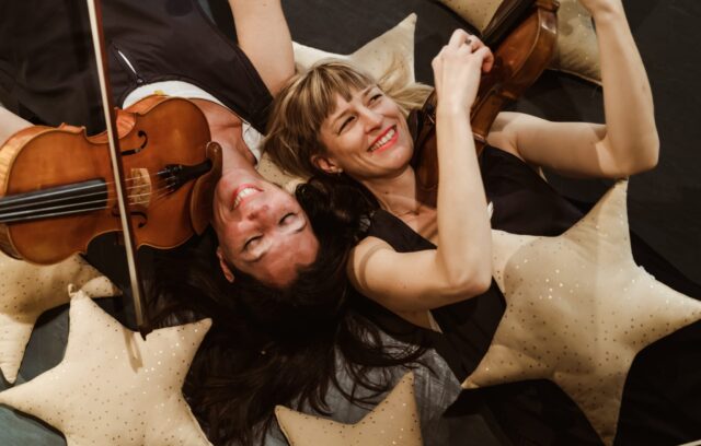 Les deux musiciennes sont allongées sur le dos. Elles sourient. Elles jouent de leur instrument, le violon et l'alto.