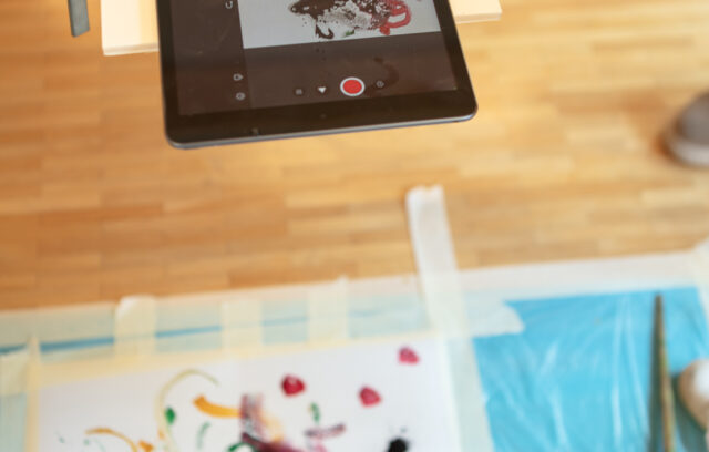 Sur l'image il y a une tablette numérique qui sert d'appareil photo. La tablette est orientée vers une feuille de papier peinte de motifs colorés. Des pinceaux et de la peinture rouge sont posés sur le côté droit.