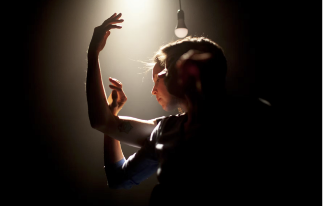 Une comédienne-danseuse se dessine en contre-jour. Elle regarde ses mains. Ses mains sont courbées vers le haut dans un geste très expressif