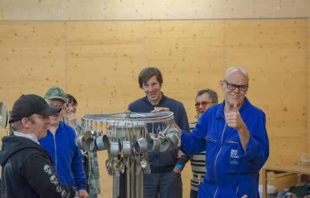 Il y a six personnes autour d'un grand instrument de musique en métal. Une des personnes porte une blouse bleue. C'est Bill Holden. Les autres personnes sont là pour jouer de la musique avec lui. Tout le monde sourit. L'instrument de musique est composé de fourchettes, de cuillères et de boîtes de conserve.