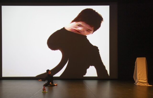 Sur une scène, une personne est à genoux face à une construction en dupplo. En fond de scène, sur le mur, sa silhouette est projetée sur un écran. La silhouette projetée est déformée en forme de vague.