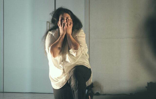Asmaa Aloui est au centre de la scène. Elle pose un genou à terre. Elle se cache le bas du visage avec les mains. Elle semble vivre une émotion très forte. Peut-être de la peur. Peut-être de la tristesse.